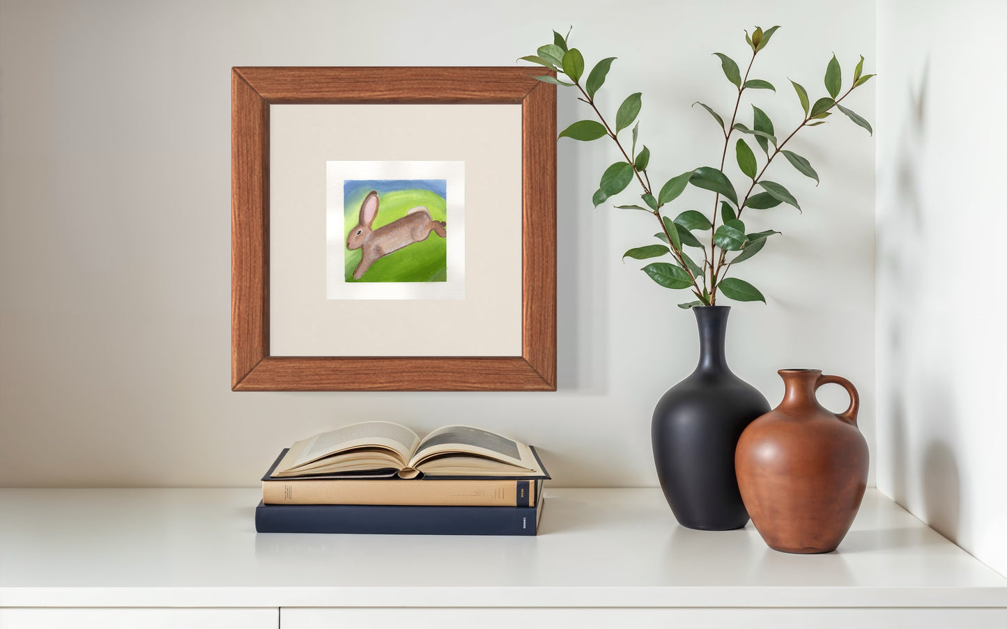 Framed artwork of a rabbit on a wall above books with two vases below.