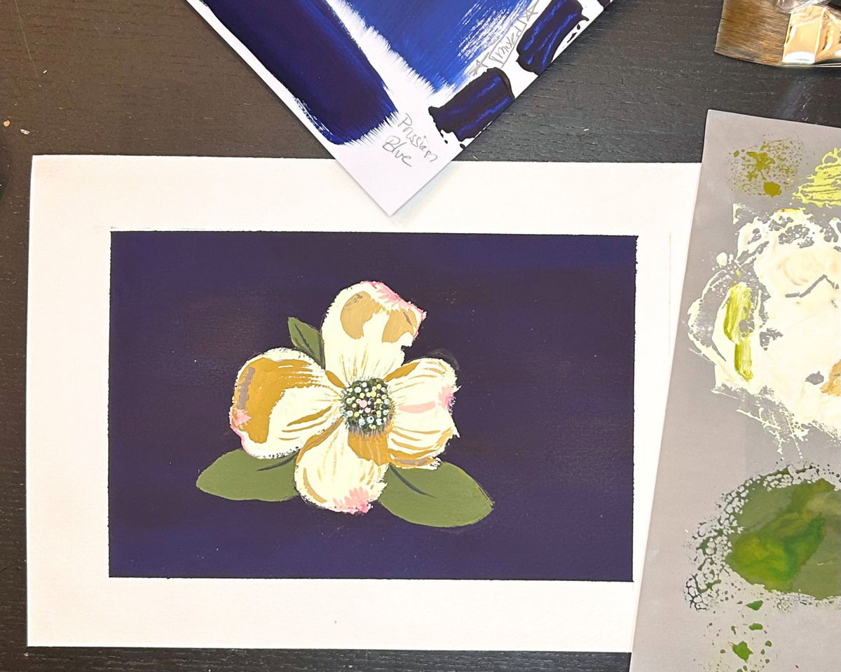 A 5x7 original painting featuring a stylized white and yellow dogwood flower with green leaves on a dark navy background, displayed on a dark tabletop next to other painted papers.