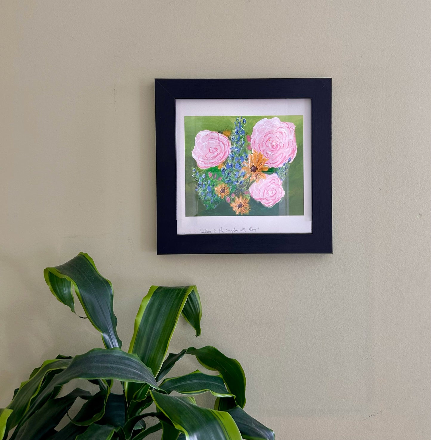 Framed artwork of pink flowers on a beige wall with a plant in the foreground.