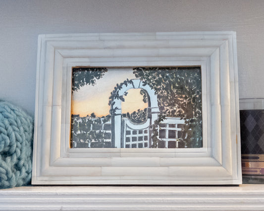 Framed mini painting on a shelf—white ridged frame around a scene of a white garden gate and trellis arch at sunset, ivy and trees in silhouette; blue knit blanket on the left and a dark candle on the right.