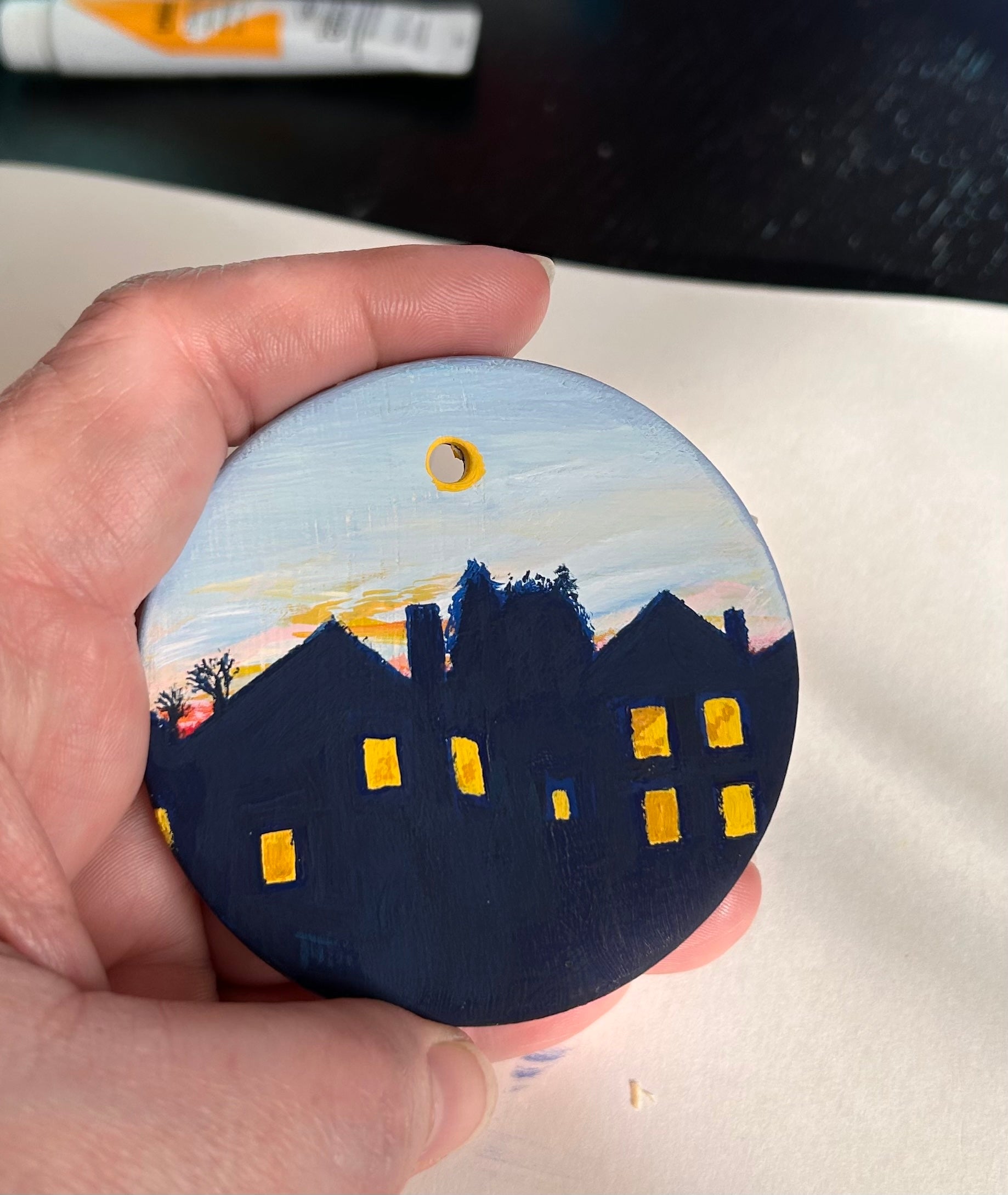 Hand holding round ceramic ornament with silhouetted houses and glowing windows under a sunset sky.  There is a tube of acrylic gouache in the background.