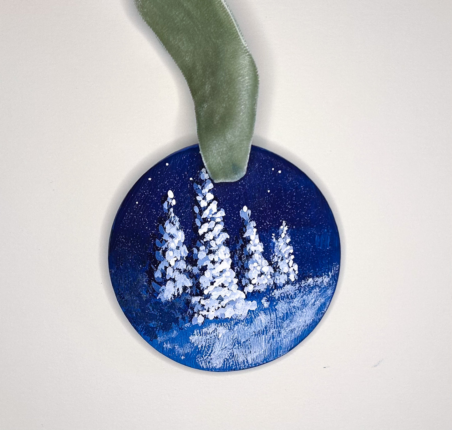 Original hand-painted round, flat ornament  of snow-covered evergreens against a starry night sky.