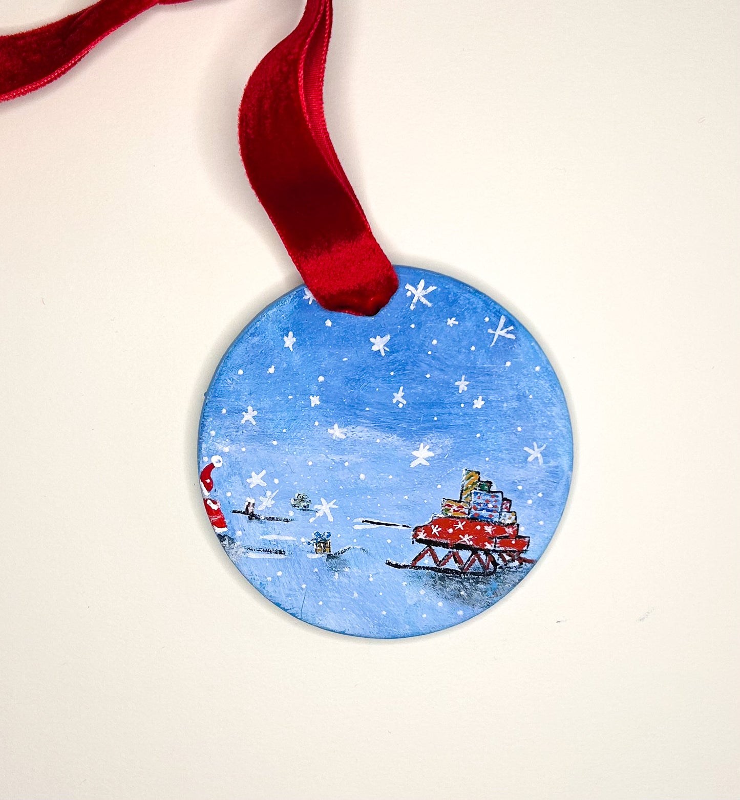 Original hand-painted circular Christmas ornament with Santa Claus and sleigh on a blue snowy background, attached to a red ribbon.