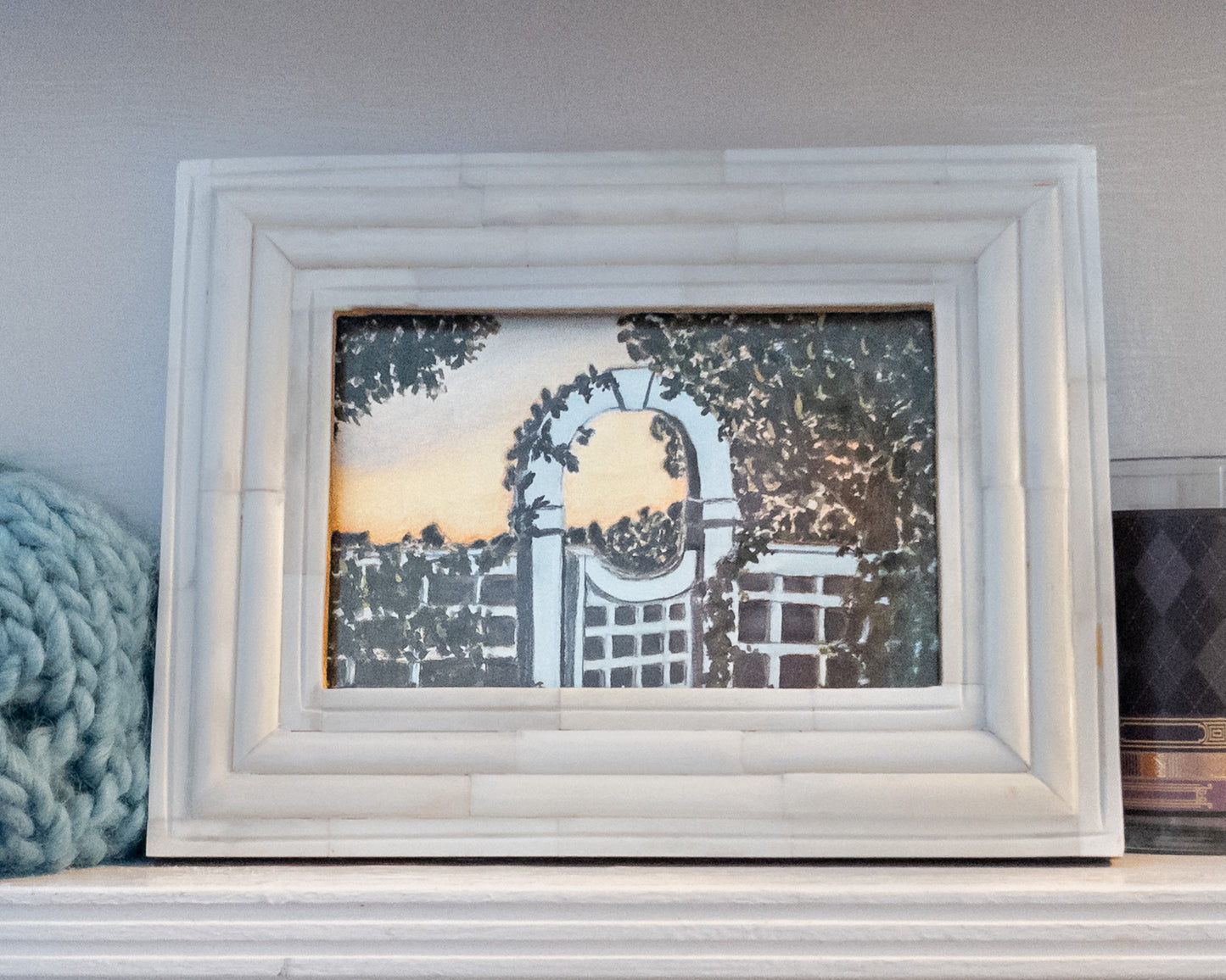 Framed mini painting on a shelf—white ridged frame around a scene of a white garden gate and trellis arch at sunset, ivy and trees in silhouette; blue knit blanket on the left and a dark candle on the right.