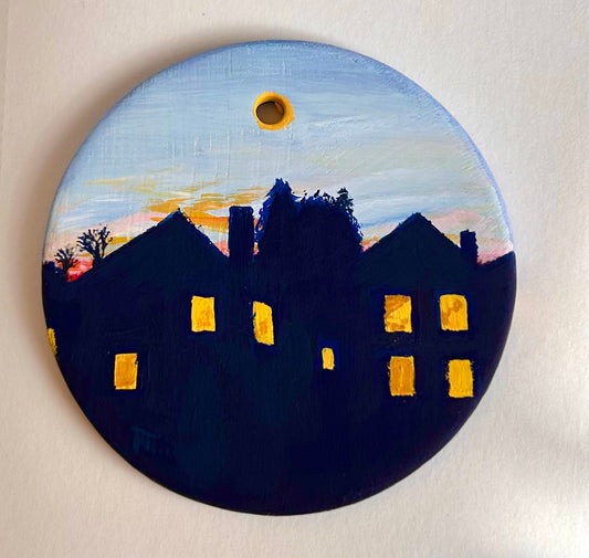 Hand-painted round ceramic ornament showing dark house silhouettes against a soft sunset sky. The windows glow warm yellow, and the sky fades from blue to peach. Painted in gouache with a small hanging hole at the top.