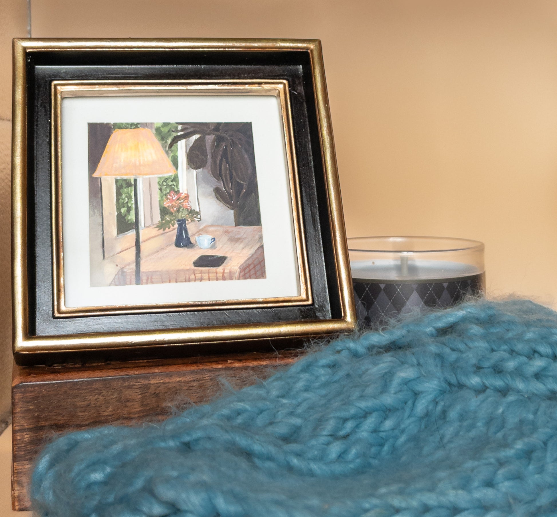 Small framed painting of a lamplit kitchen table on a wooden block beside a gray candle and a blue knit blanket; neutral peach wall behind.