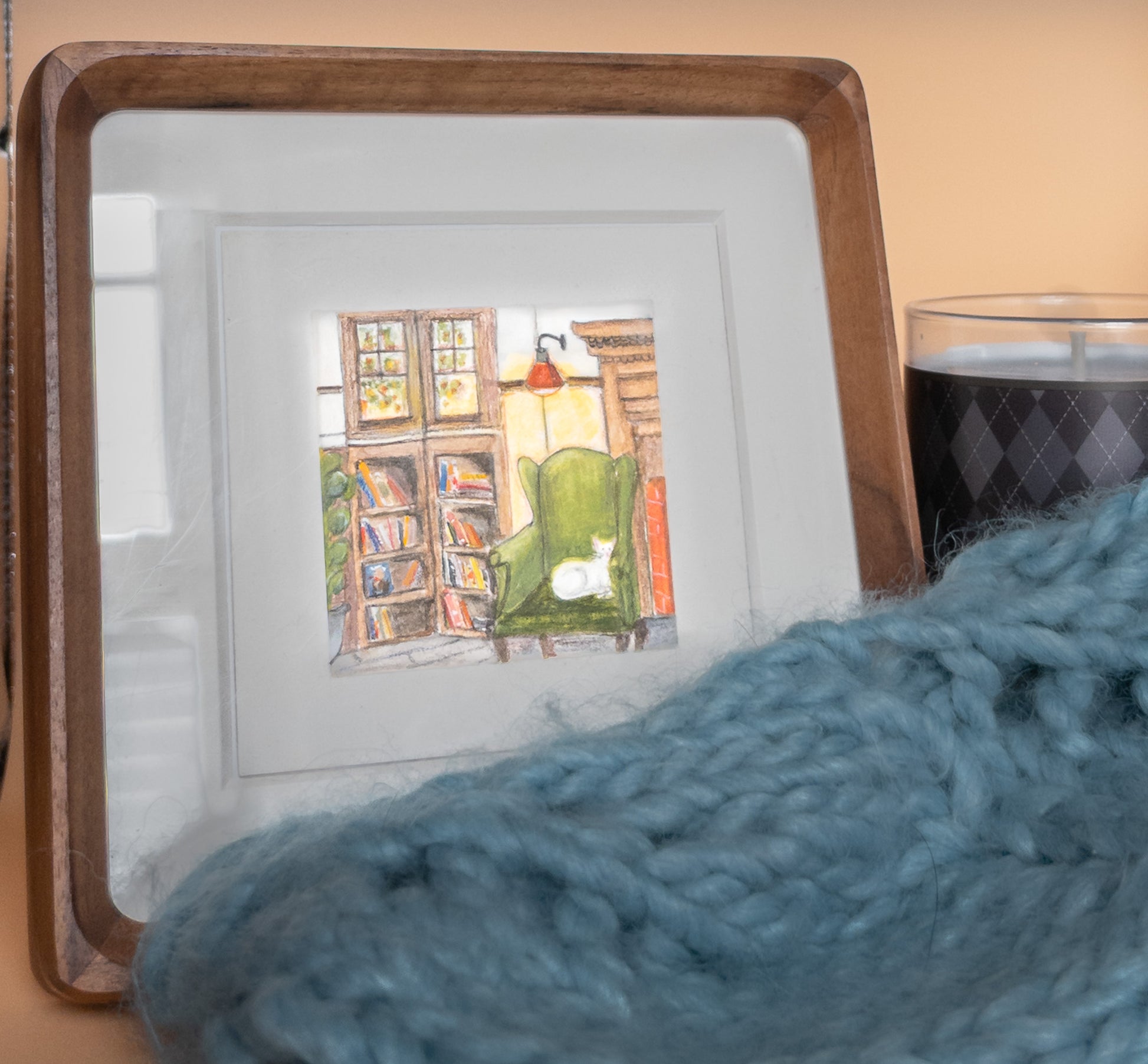 Framed mini painting of a cozy library corner—green chair, bookshelves, white cat—leaning on a shelf beside a dark candle, with a blue knit blanket in front and a peach wall behind.