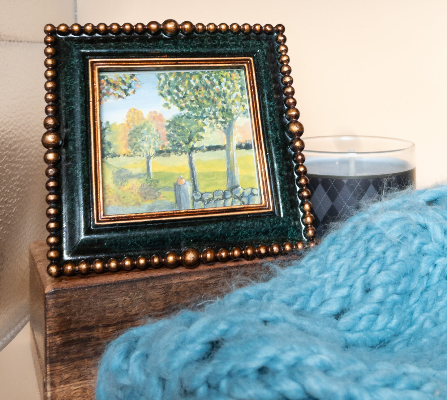 Small framed landscape painting in a dark green beaded frame, resting on a wooden block beside a gray candle, with a blue knit blanket in the foreground and a peach wall behind.